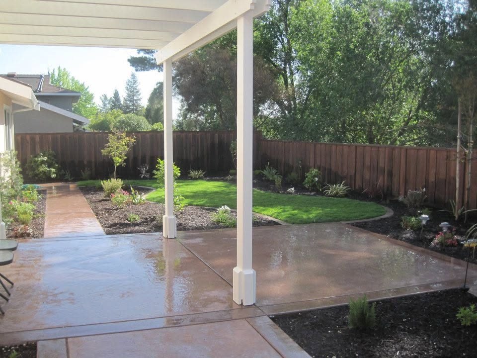 Colored concrete patio beneath a white patio cover beside a lawn and wood fence