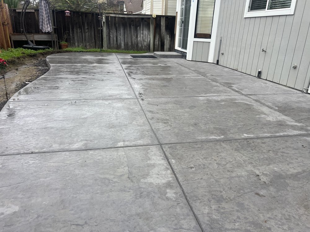 Large light gray broom finish concrete patio beside a house and wood fence