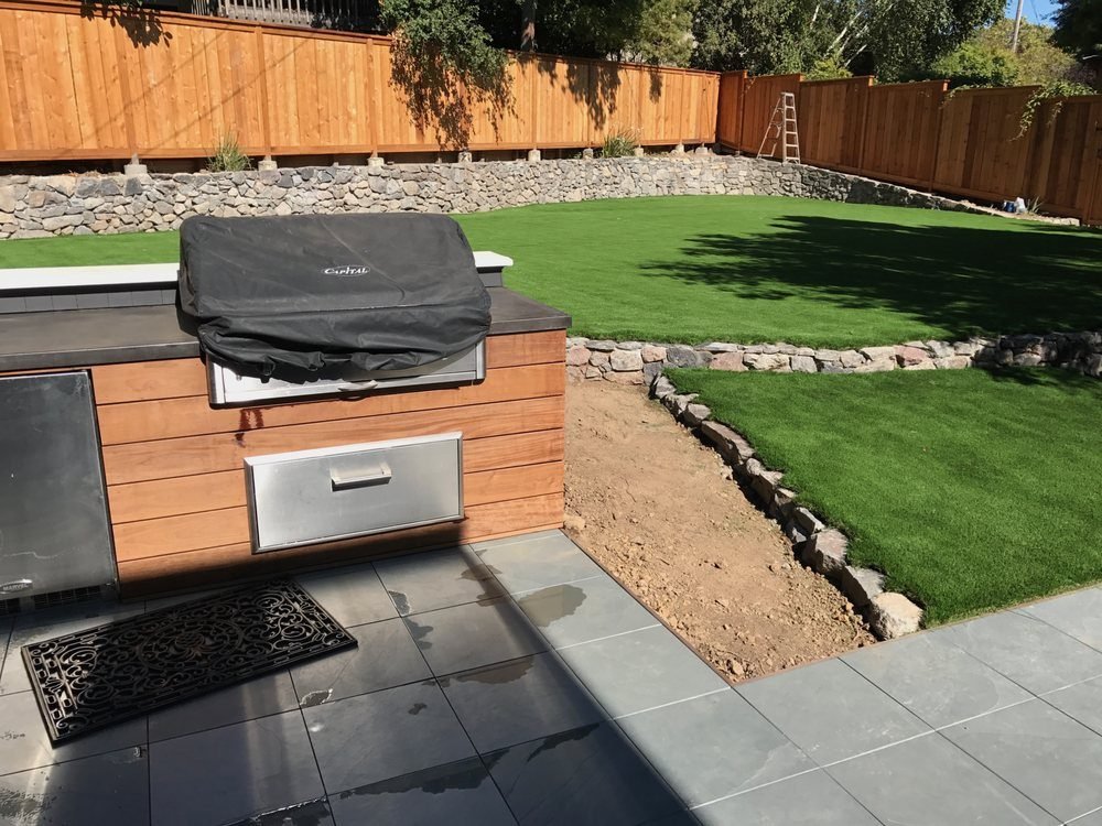 Built-in BBQ island with a covered grill and metal drawers beside a turf lawn and stone retaining wall